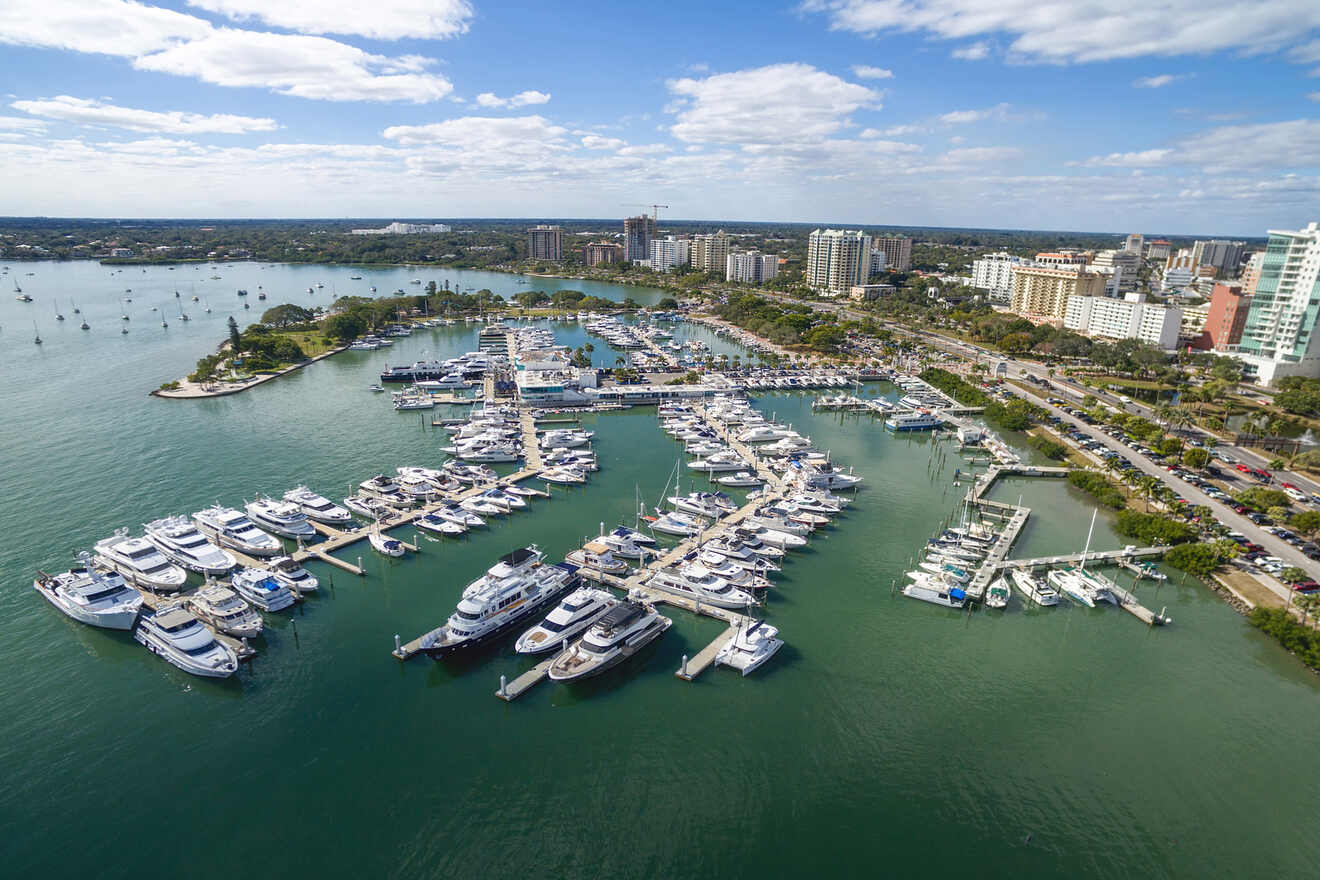 An aerial view of a marina filled with yachts and boats, with a coastal city skyline and calm waters in the background under a blue sky with scattered clouds.