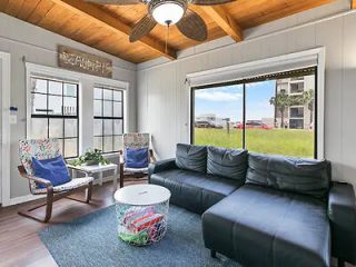 A cozy living room with a black sectional sofa, colorful chairs, a round white table, and a beach-themed decor. Large windows offer a view of a grassy area and nearby buildings. Ceiling fan above.