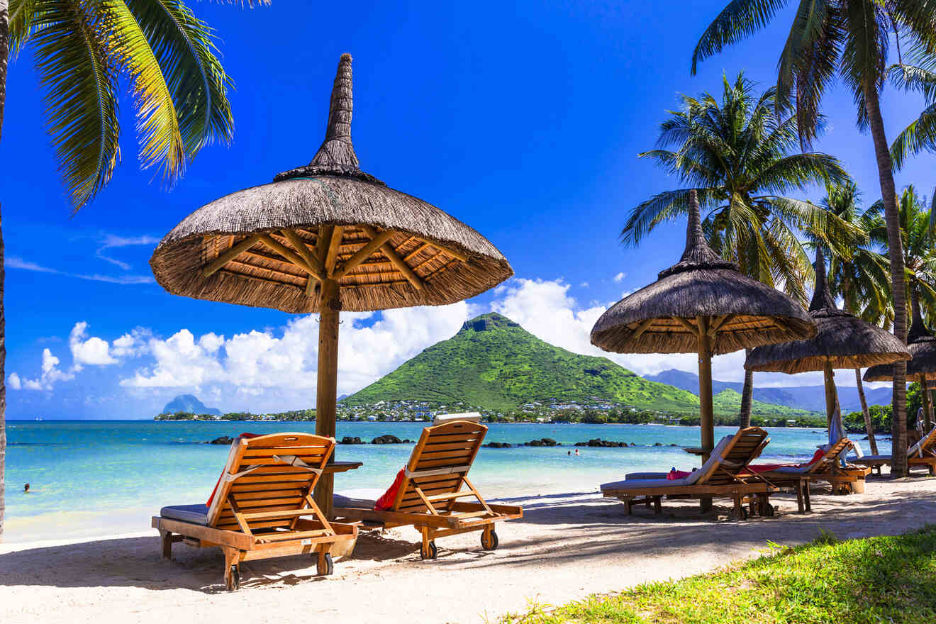 Resort beachfront view featuring thatched umbrellas and wooden loungers facing a majestic mountain across the calm sea, conveying a serene tropical paradise vibe