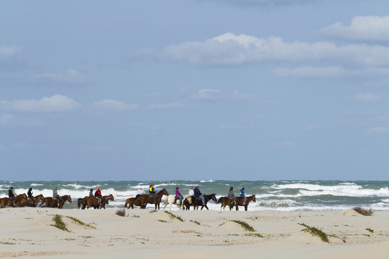A group of people riding horses along a sandy beach with the ocean waves and blue sky with clouds in the background.