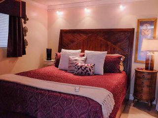 A bedroom with a large bed featuring a dark red bedspread and multiple pillows, a wooden headboard, a bedside table with a lamp, and wall-mounted lights above the bed.