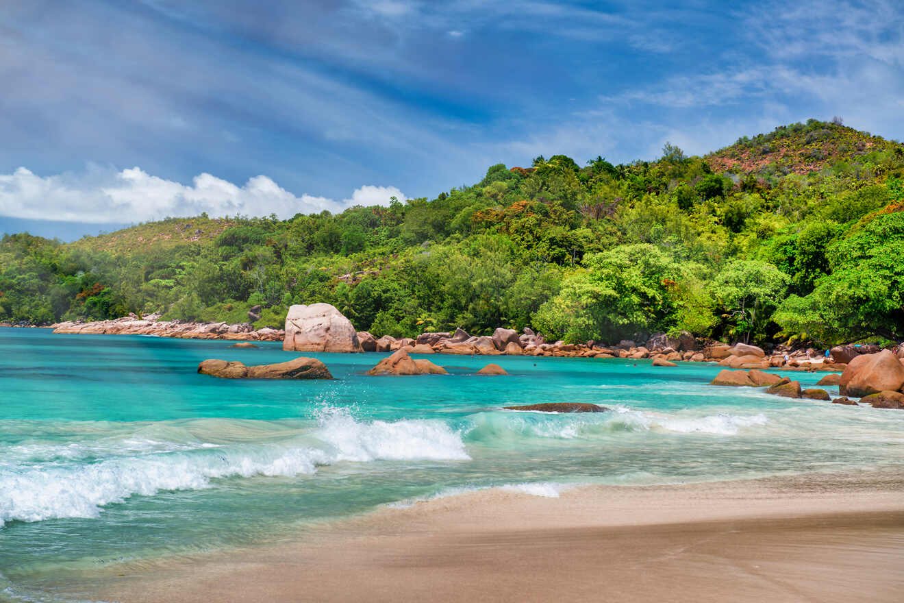 A picturesque beach scene with turquoise water, gentle waves, and large rocks along the shore, framed by lush green hills.