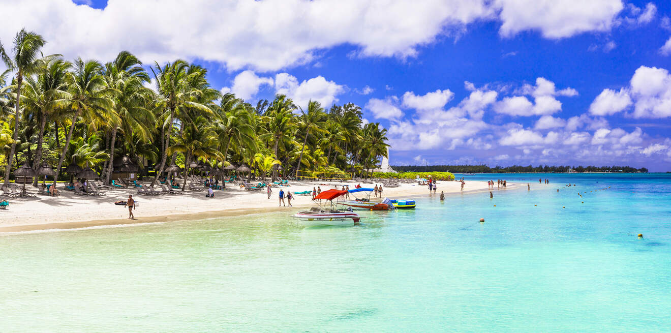 A picturesque beach scene with white sand, crystal clear waters, and a row of palm trees, where visitors enjoy various beach activities and boat rides under a partly cloudy sky