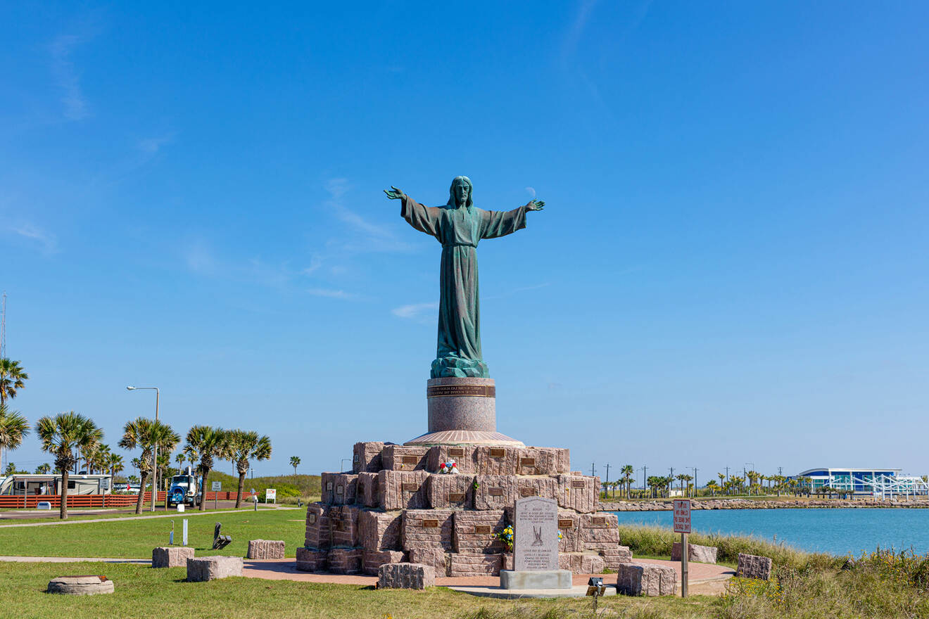 A statue of Jesus with outstretched arms stands atop a stone base surrounded by grass, with a blue sky and waterfront in the background. Palm trees and a few buildings are visible in the distance.