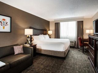 A hotel room with a large bed, a sofa, bedside lamps, a desk with a red chair, and a TV. A window with curtains is in the background.