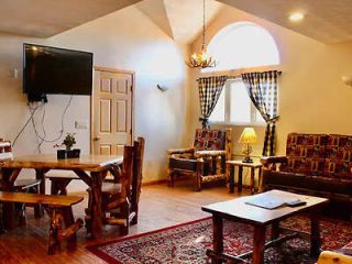 Cozy living room with wooden furniture, mounted TV, window with floral curtains, two chairs, and a table on a patterned rug. A door and a ceiling light are visible.