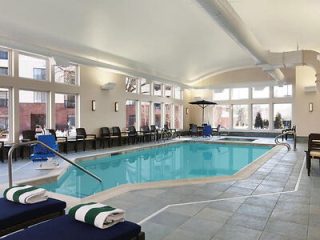 Indoor swimming pool area with lounge chairs, towels, and bright lighting. Large windows line one wall, and a pool lift is visible by the poolside.