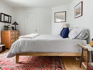 A tidy bedroom with a bed, gray bedding, two pillows, a wooden dresser, and a side table. Two framed pictures are on the walls, and a red patterned rug covers the floor.