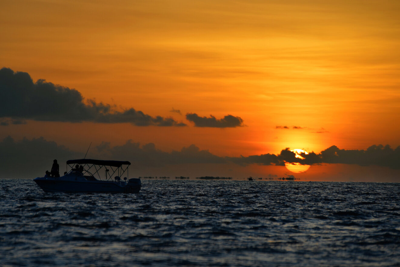 A boat floats on the water during a vibrant orange sunset, with clouds dotting the sky above the horizon.