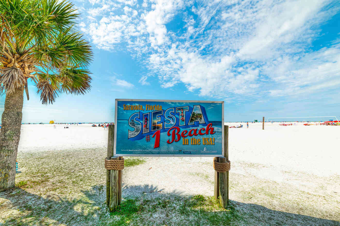 A large sign that reads "Sarasota, Florida: Siesta #1 Beach in the USA" standing on the white sand of a sunny beach, with palm trees and people in the background.