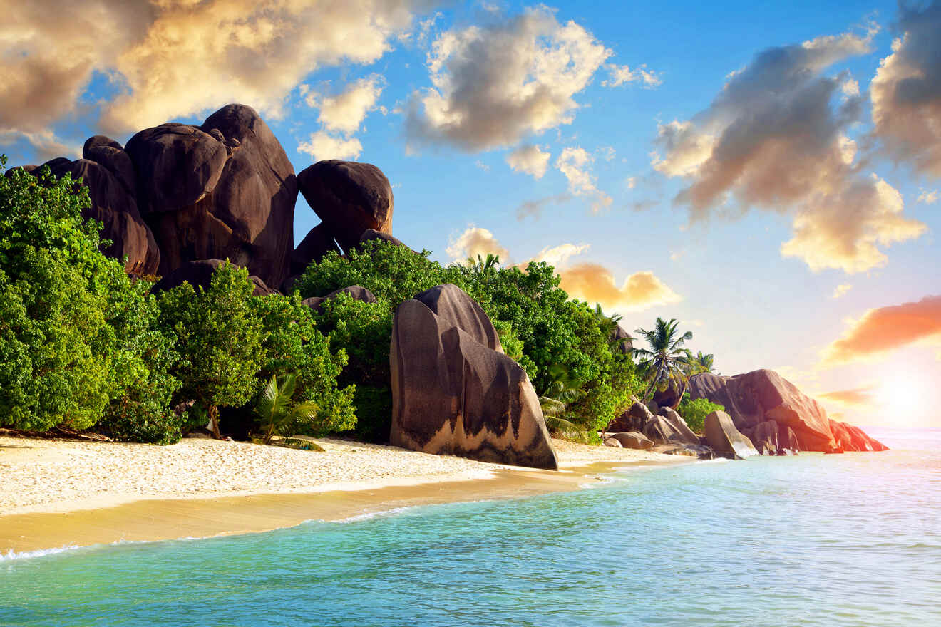 A scenic beach at sunset, with large boulders, lush greenery, and the sun setting over the water, casting a warm glow.