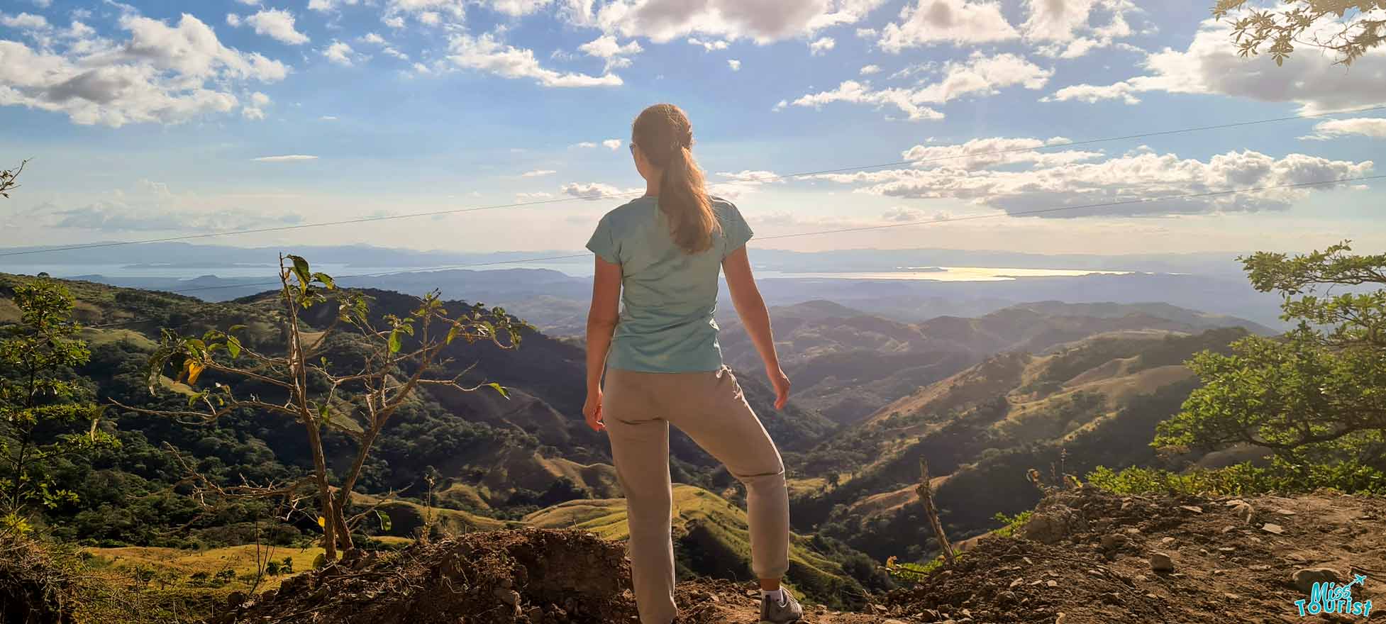 Author of the post stands on a hilltop, facing away, overlooking a scenic landscape with rolling hills, a body of water, and a partly cloudy sky.