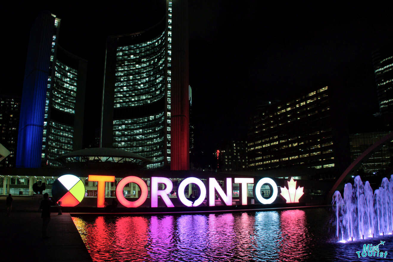 Nighttime view of Toronto sign illuminated in vibrant colors with the reflection in a fountain at Nathan Phillips Square, flanked by the curved towers of Toronto City Hall