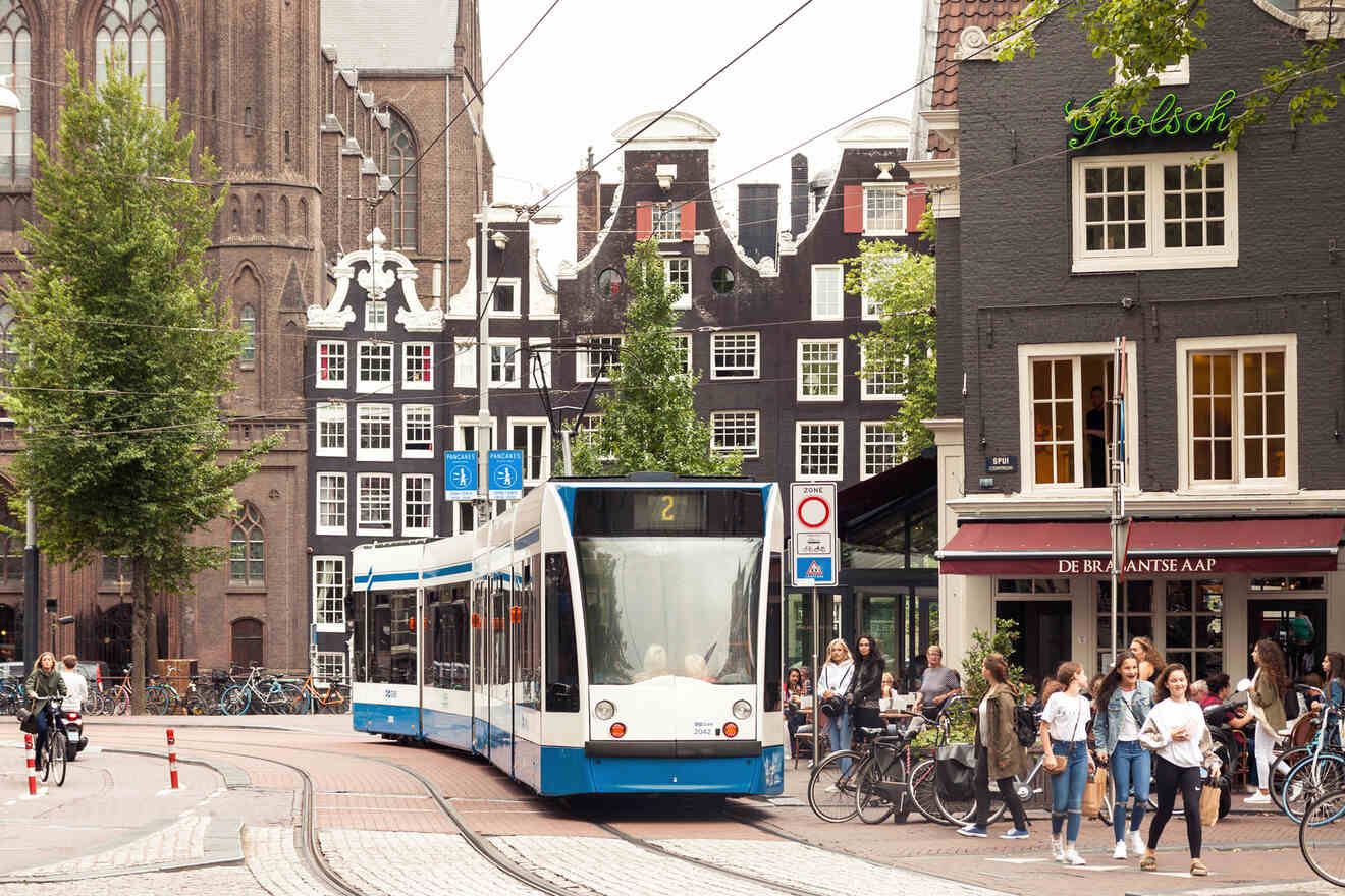 A blue and white tram travels through a busy intersection in an urban area with historic buildings and people walking and biking around.