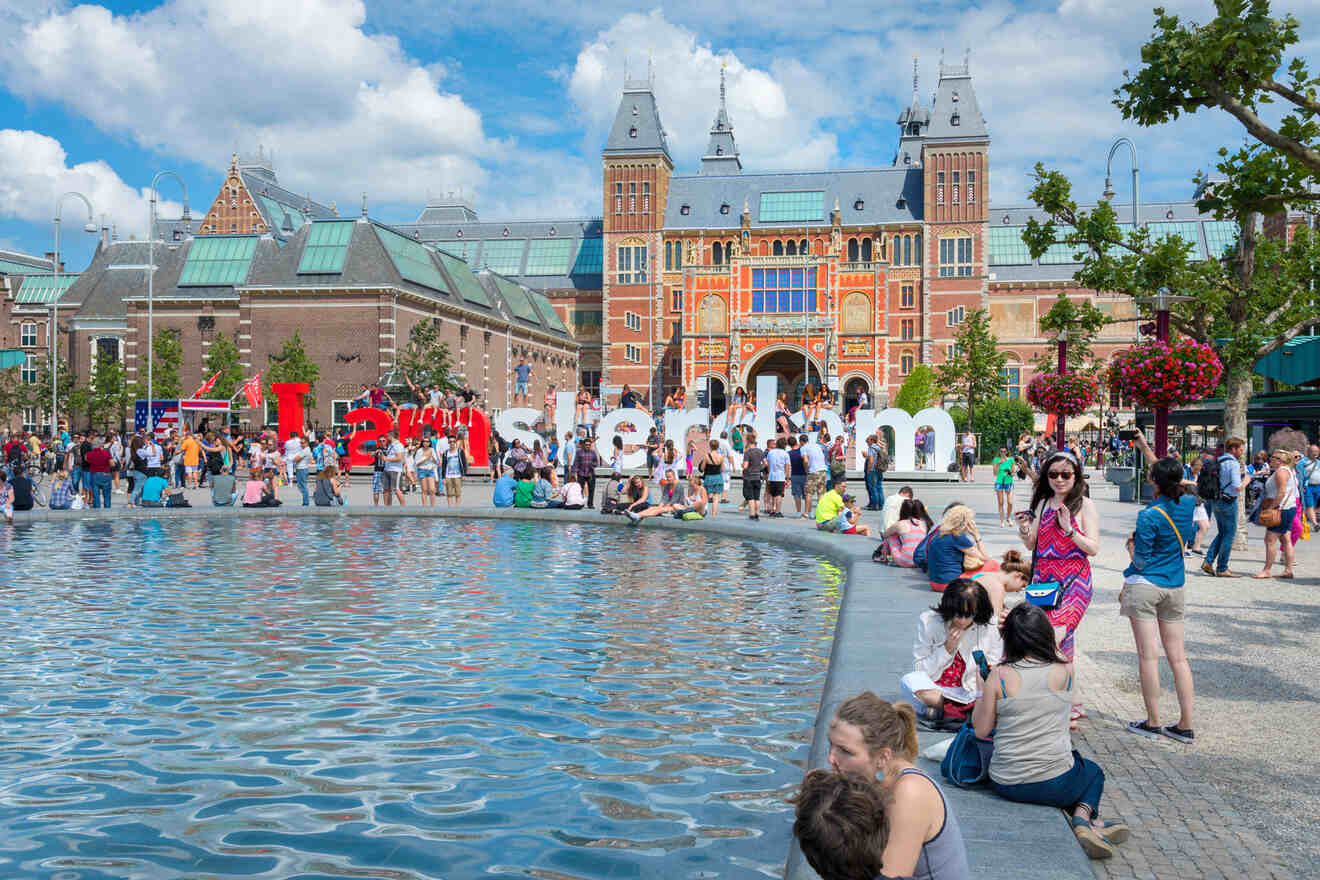 People gather around a reflective pool with the Rijksmuseum and large "I amsterdam" sign in the background.