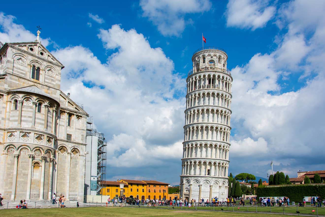 View of the Leaning Tower of Pisa and the adjacent Pisa Cathedral under a partly cloudy sky, with tourists on the grass and pathways surrounding the historic buildings.