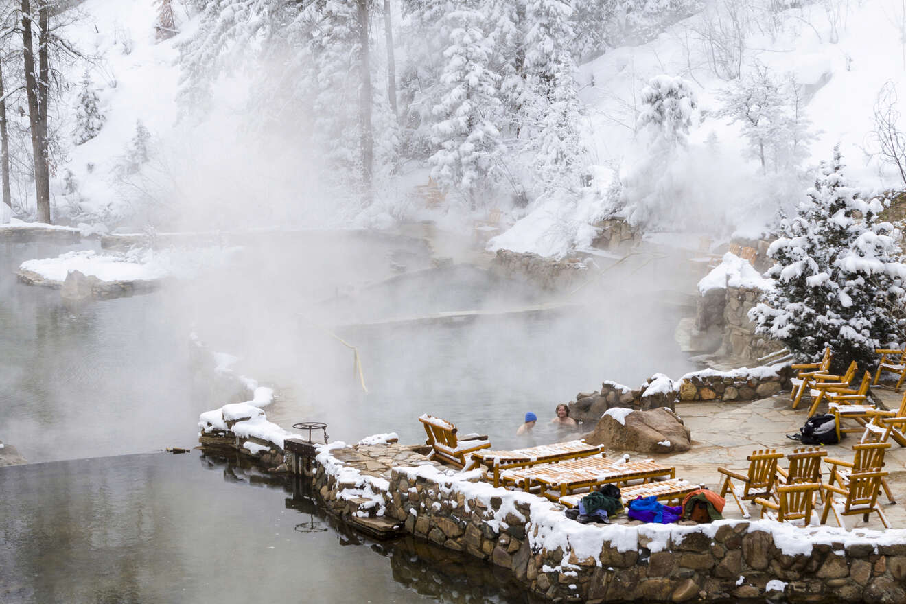 People relaxing in a hot spring surrounded by snow-covered trees and rocks; wooden chairs and backpacks are nearby.