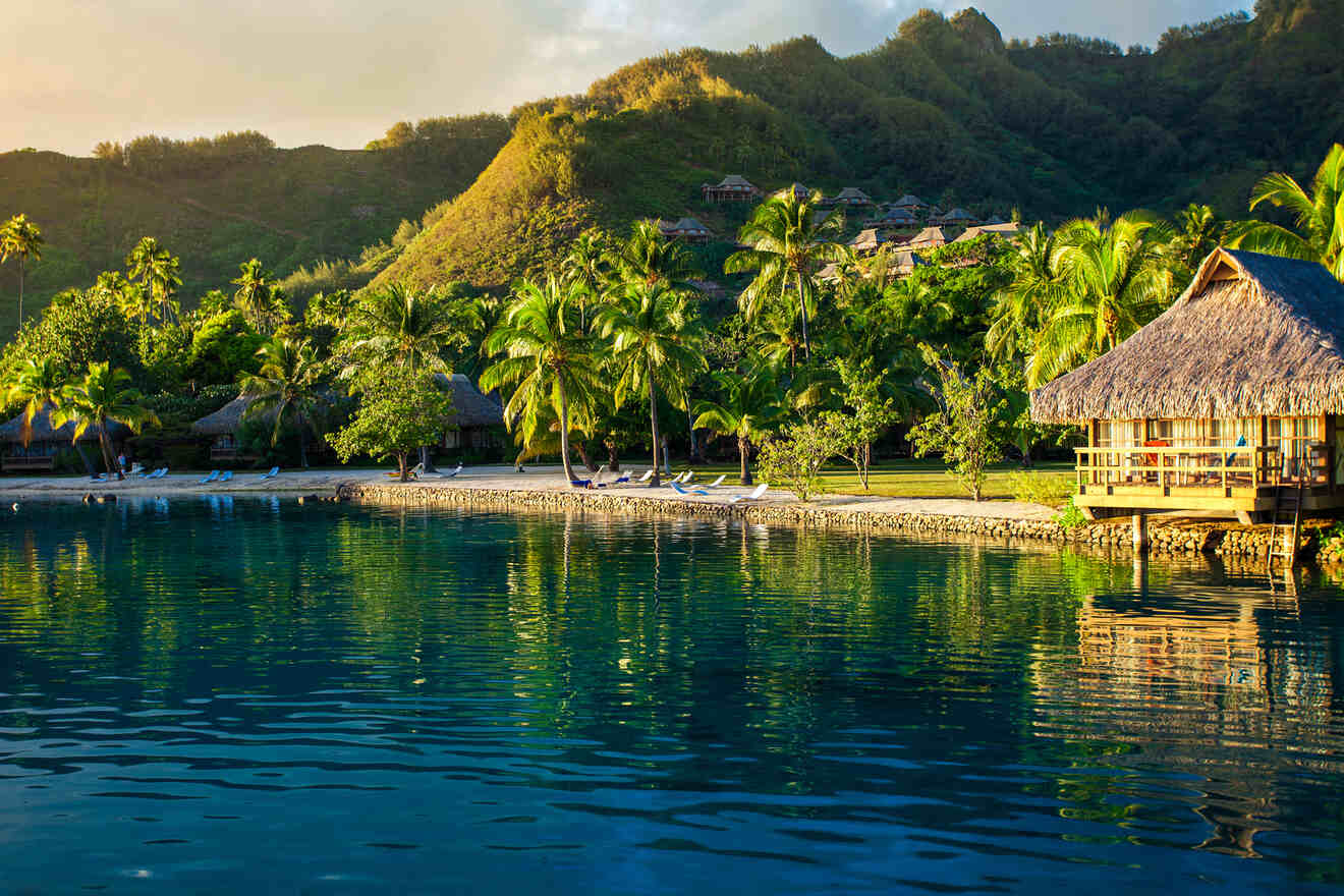 An overwater bungalow where to stay in Moorea with dark blue water, next to a palm tree-lined beach with a mountain background. 