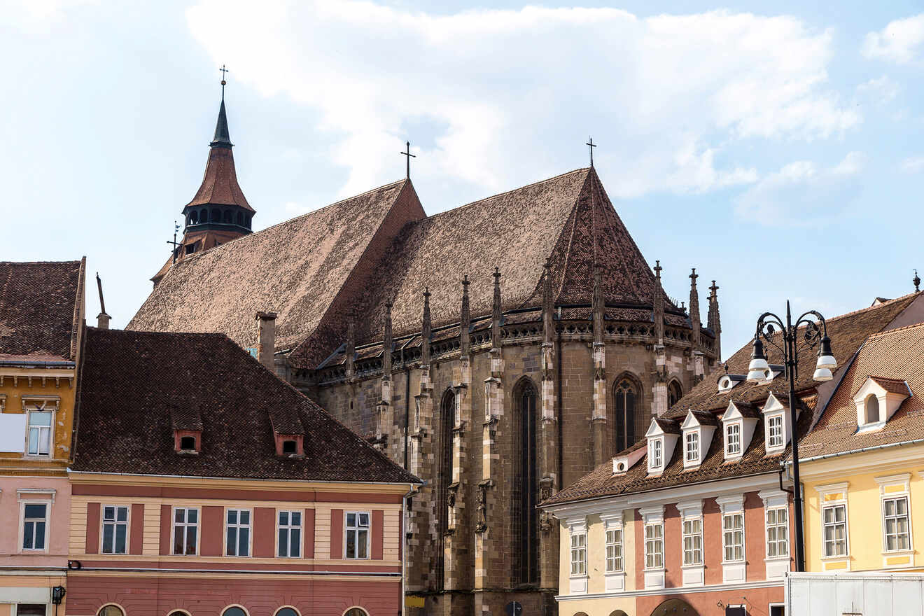 A large, historic church with Gothic architecture is surrounded by colorful buildings under a partly cloudy sky.