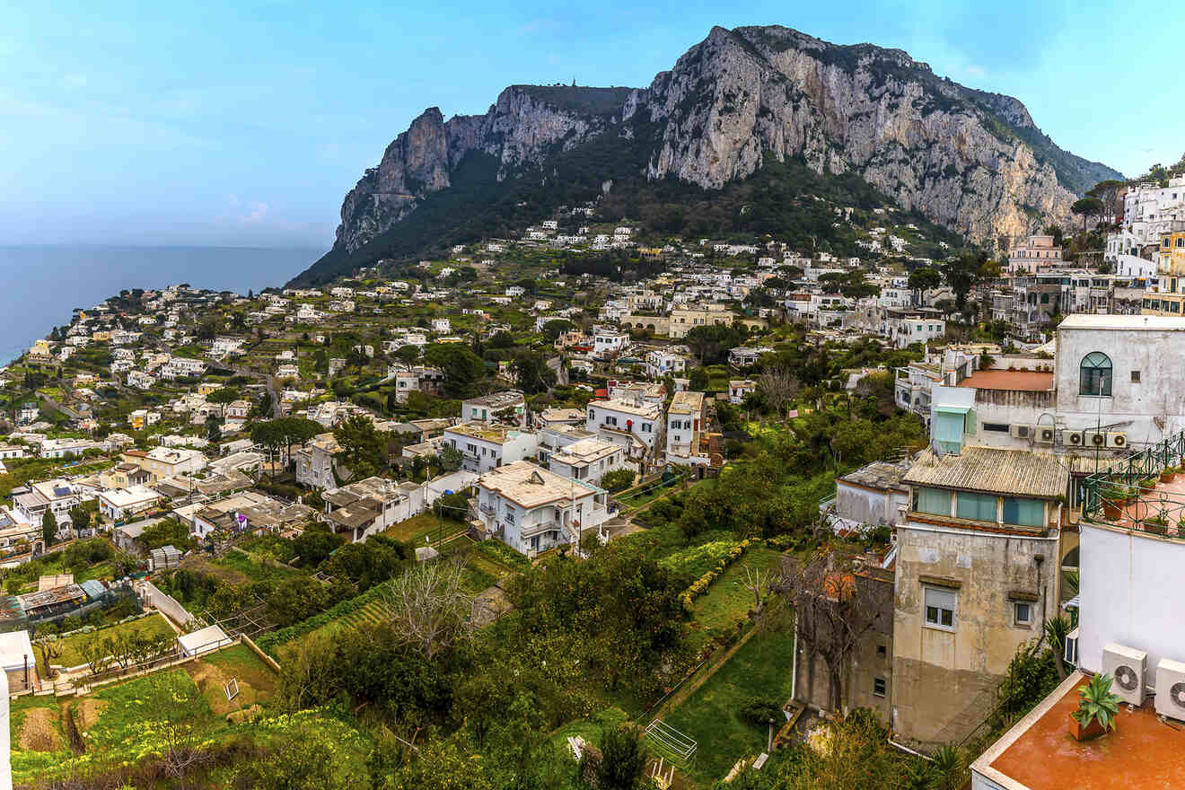 A scenic view of a hillside town with white buildings, greenery, and a rocky mountain in the background under a clear sky.