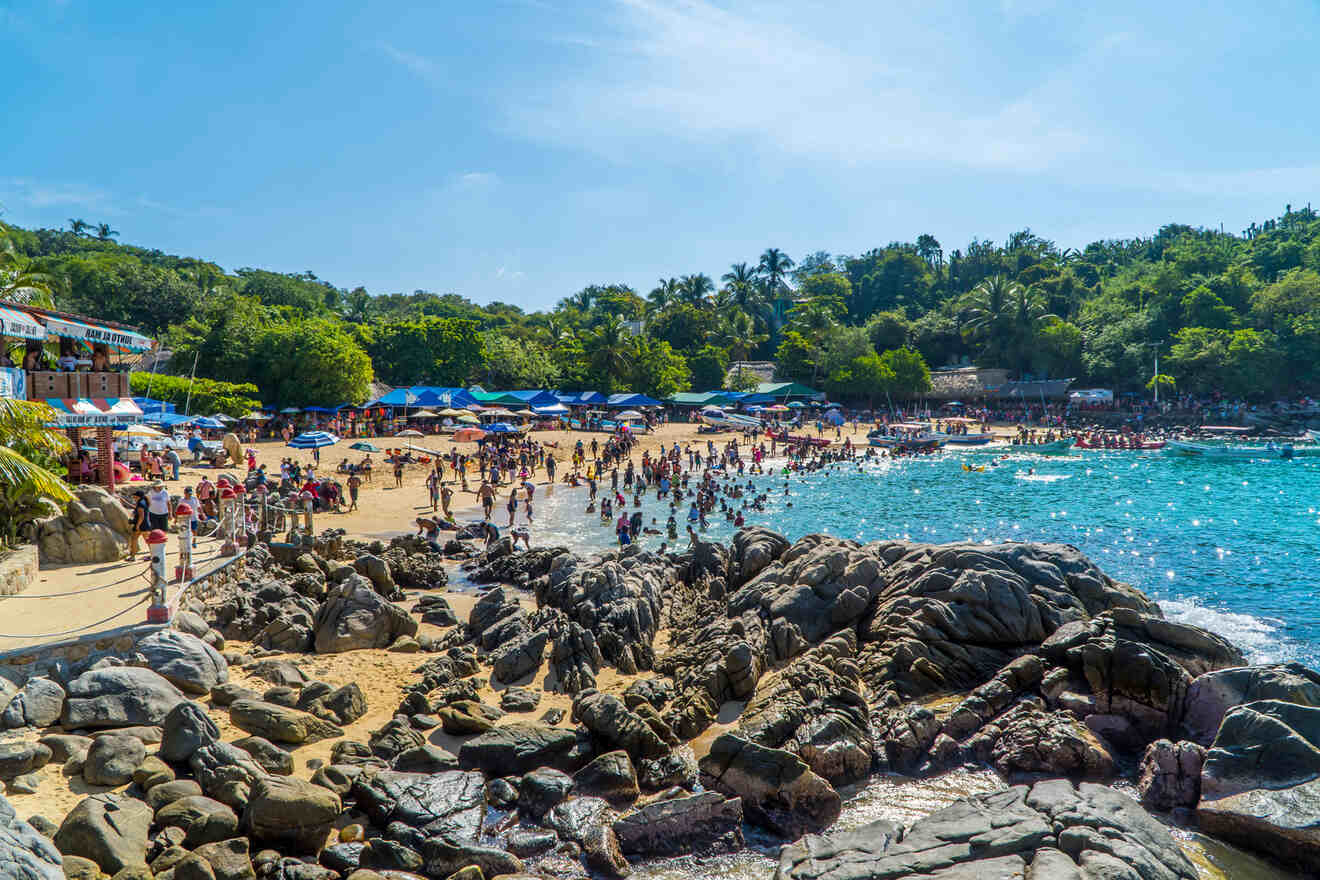 A view of rocks, ocean, and a Playa Puerto Angelito beach full of people. One of the best things to do in Puerto Escondido