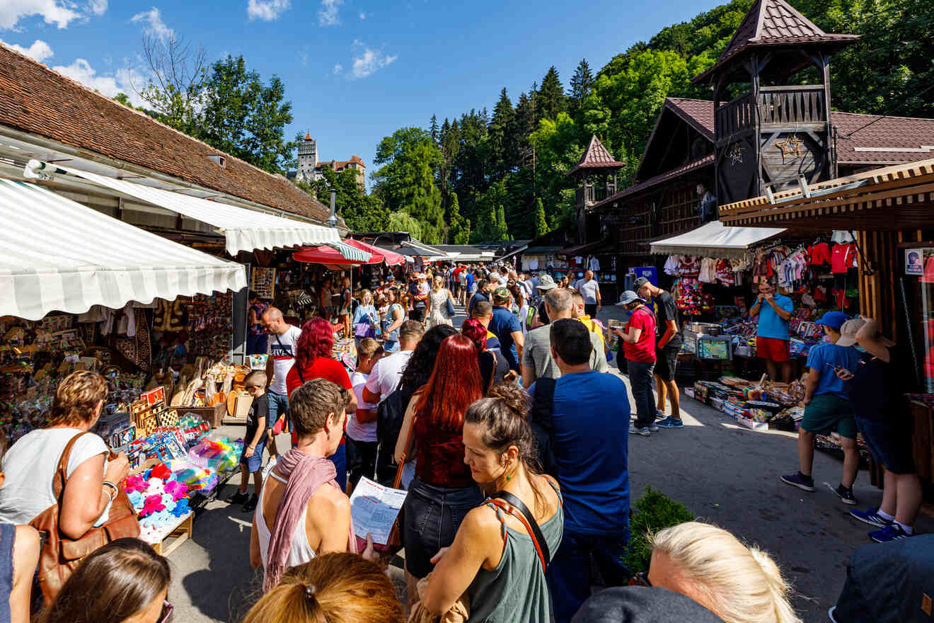 A bustling outdoor market with numerous shoppers exploring various stalls selling souvenirs, crafts, and clothing under sunny weather. Trees and rustic buildings surround the scene.