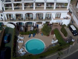 Aerial view of a hotel with two floors of balconies, an outdoor pool surrounded by lounge chairs, and several parked cars nearby.