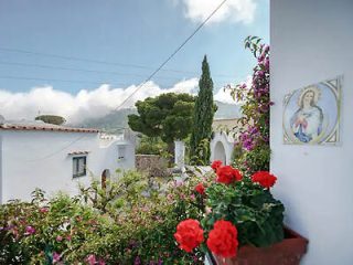 A quaint village scene with white buildings and colorful flowers on a balcony. Hills and trees are visible in the distance under a partly cloudy sky. A small religious icon is mounted on a wall.
