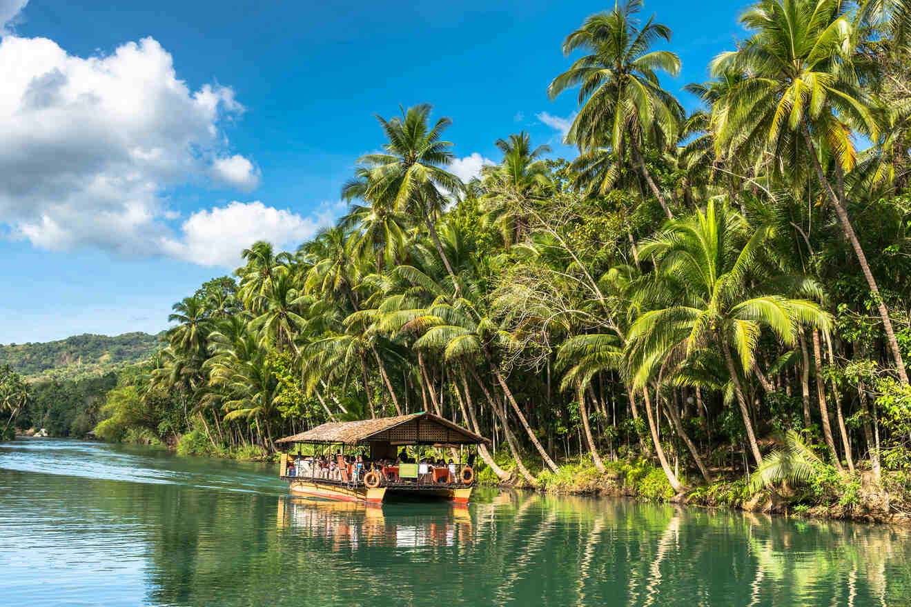 An image of a raft with roof floating down a river with lush palm trees by the side, the Loboc River Cruise is one of the must-visit attractions on your 3 day Bohol itinerary