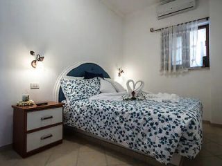 A neatly made bed with floral bedding and towel swans on top, situated in a white-walled bedroom with wall-mounted lights, a small window with a curtain, and a bedside table.