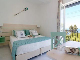 A bright bedroom with a double bed decorated with teal accents, a wall-mounted paddle, an open glass door revealing a balcony with a sea view, and a small table with a bottle and glass.