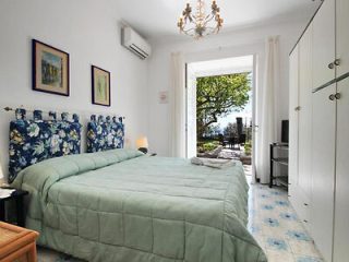 A bedroom with a green comforter on a double bed, white walls, two framed pictures, air conditioning, and a view of an outdoor terrace through double doors.