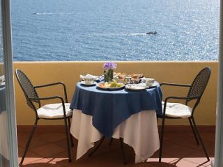 A round table with a blue and white tablecloth, set for two, on a balcony overlooking the ocean. The table has a vase with purple flowers, plates, cups, and food. Two chairs are placed around the table.