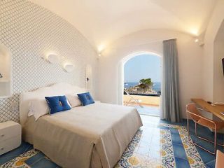 A bedroom with a double bed, blue and white patterned floor tiles, a desk, and an open door leading to a balcony with an ocean view. The room has white walls and is decorated with blue accents.