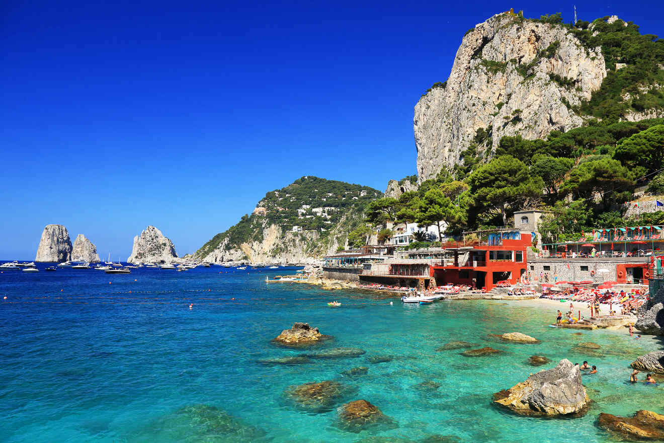 Coastal scene with clear turquoise water, rocky shoreline, and buildings built into a cliffside. Several boats are anchored in the distance, and people are sunbathing and swimming near the shore.