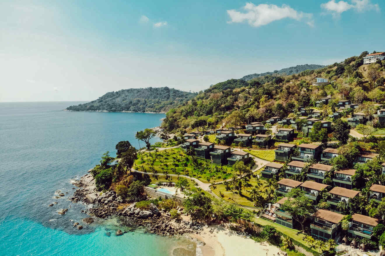 Aerial view of hillside villas surrounded by lush greenery overlooking a turquoise ocean and a small beach.