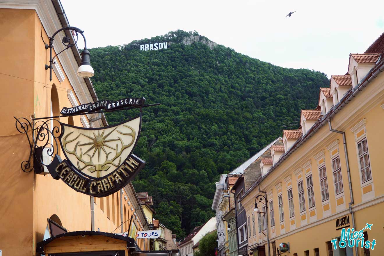 Street view of historical buildings in Brasov, Romania, with a forest-covered hill in the background, and a BRASOV sign on the hilltop mimicking the Hollywood sign.