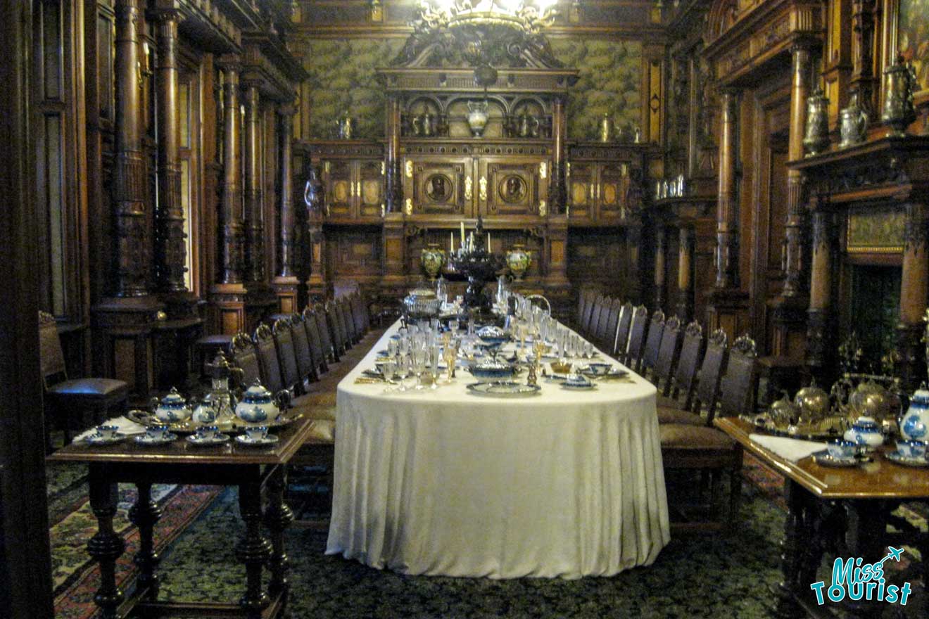 Ornate dining room with a long table set with fine china, surrounded by wooden chairs. Elaborate wood paneling and decor adorn the walls. Chandelier overhead.