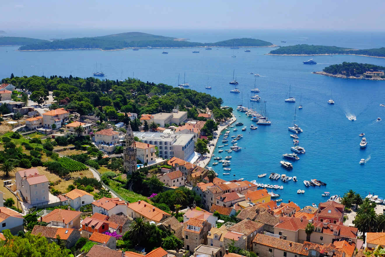 Aerial view of a coastal town with boats in the harbor, red-roofed buildings, greenery, and surrounding blue waters with small islands in the distance.