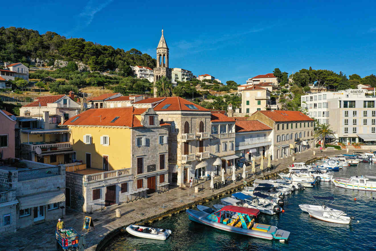 A waterfront scene in a coastal town featuring boats docked at a marina, a line of colorful buildings, and a bell tower in the background against a hillside of green trees under a blue sky.