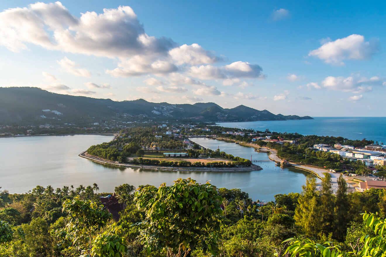 Aerial view of a peninsula with buildings and lush greenery, surrounded by water, under a blue sky with scattered clouds, and mountains in the background.
