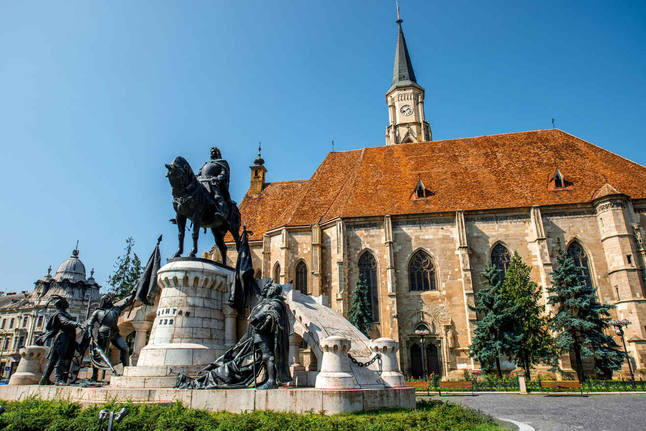 Statue of a horse and rider in front of a historic church with a tall, pointed tower and red-tiled roof, framed by greenery under a clear blue sky.