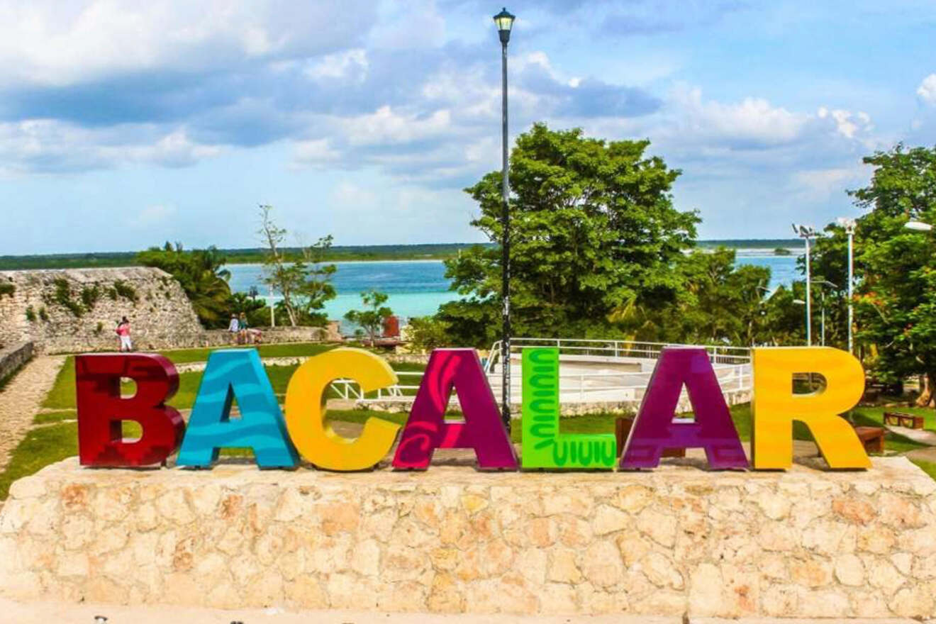 Colorful sculptures of letters spelling "BACALAR" stand on a stone platform with trees, a distant waterfront, and a partly cloudy sky in the background.