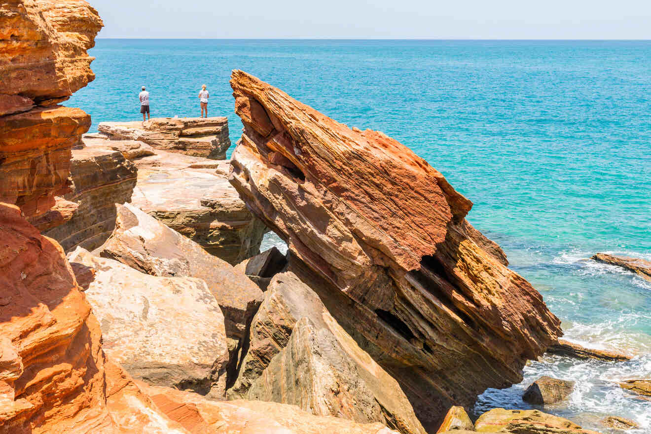 Two people standing on rocky cliffs by the ocean, with large red rocks in the foreground and blue water stretching into the horizon.