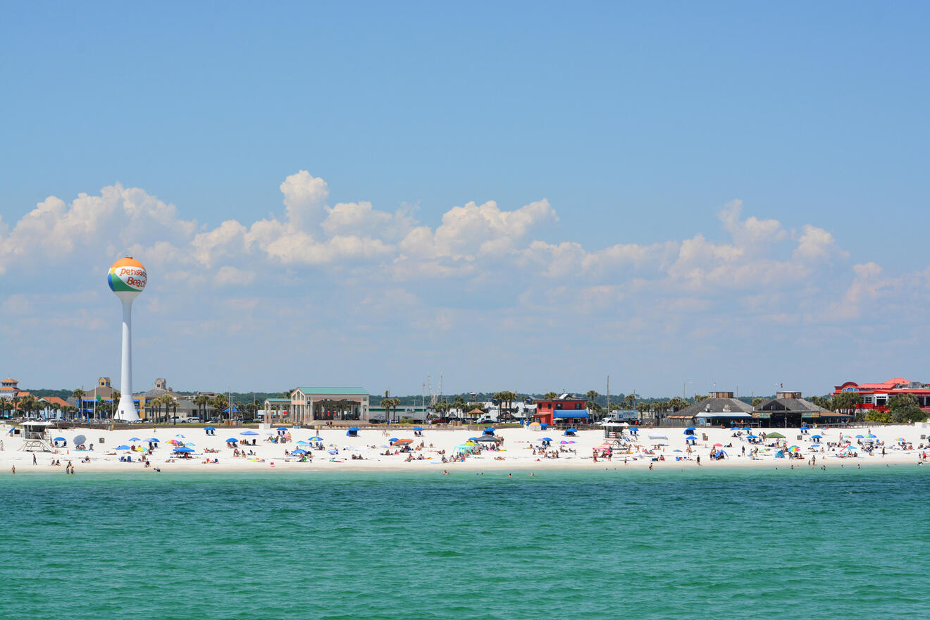 A beach scene with many people and umbrellas on white sand. A water tower and buildings are visible in the background under a partly cloudy sky.