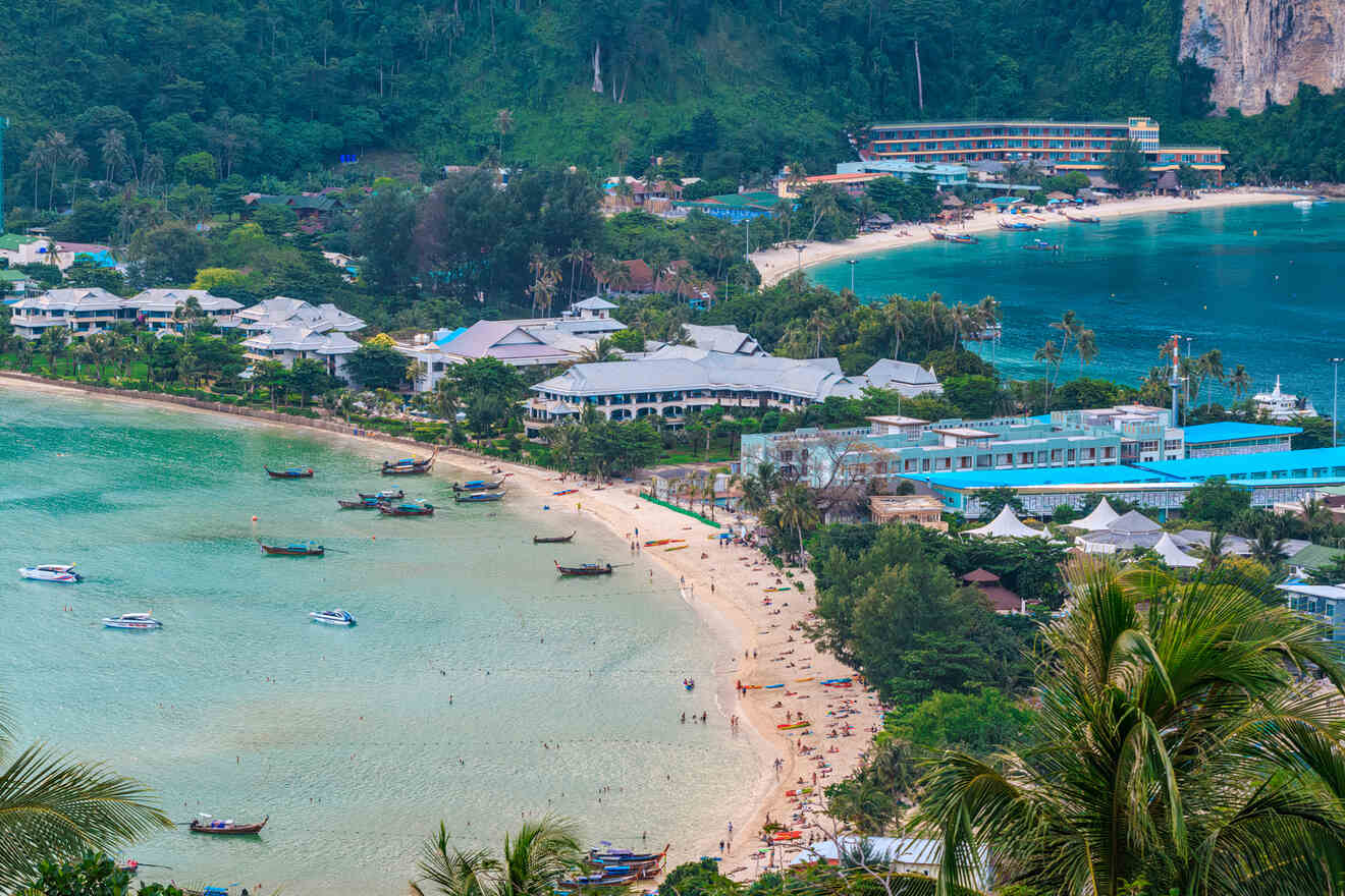 Aerial view of a bustling beach resort area with clusters of buildings, vibrant beach activity, and numerous boats in a calm bay surrounded by lush hills