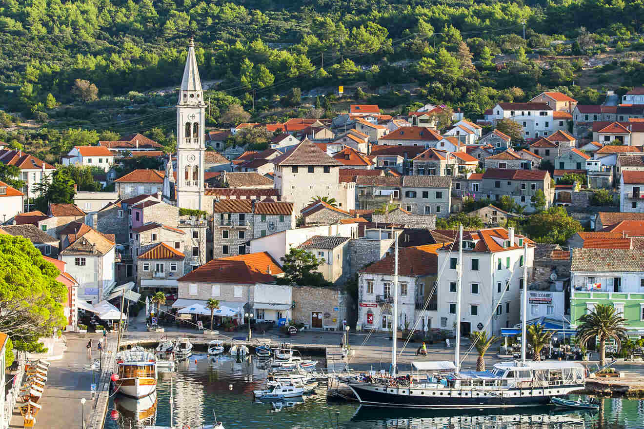 An aerial view of a coastal town with buildings featuring red roofs, a tall clock tower, boats docked in a marina, and a densely forested hillside in the background.