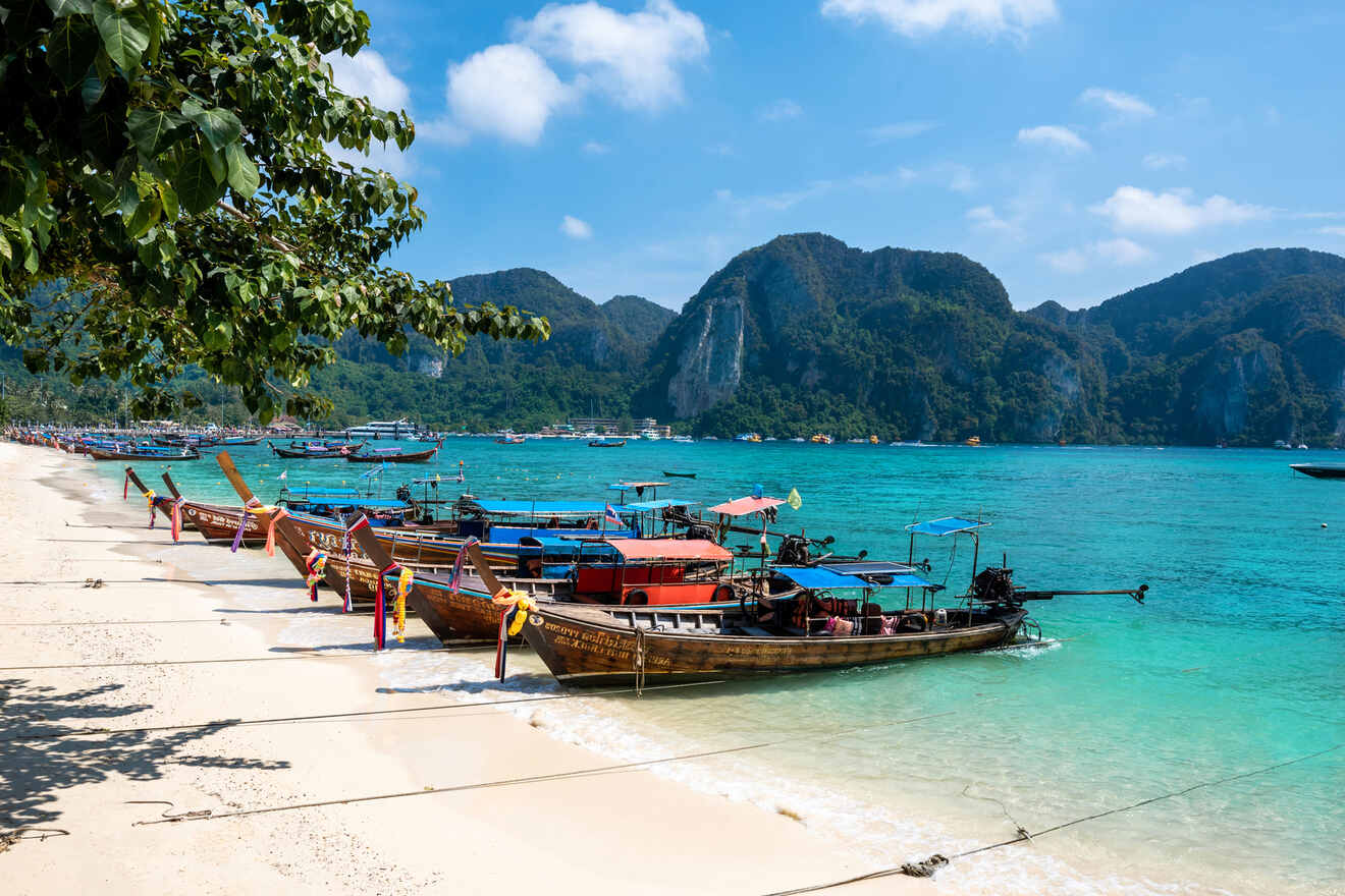 Wooden boats are anchored on a sandy beach with clear turquoise water and distant green hills under a sunny sky.