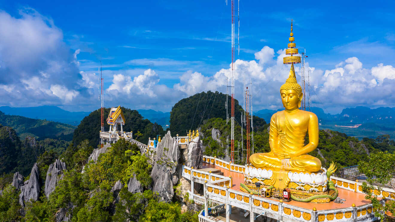 Majestic golden Buddha statue sitting atop a mountain, with a temple structure below and panoramic views of lush forests and mountain ranges in the background