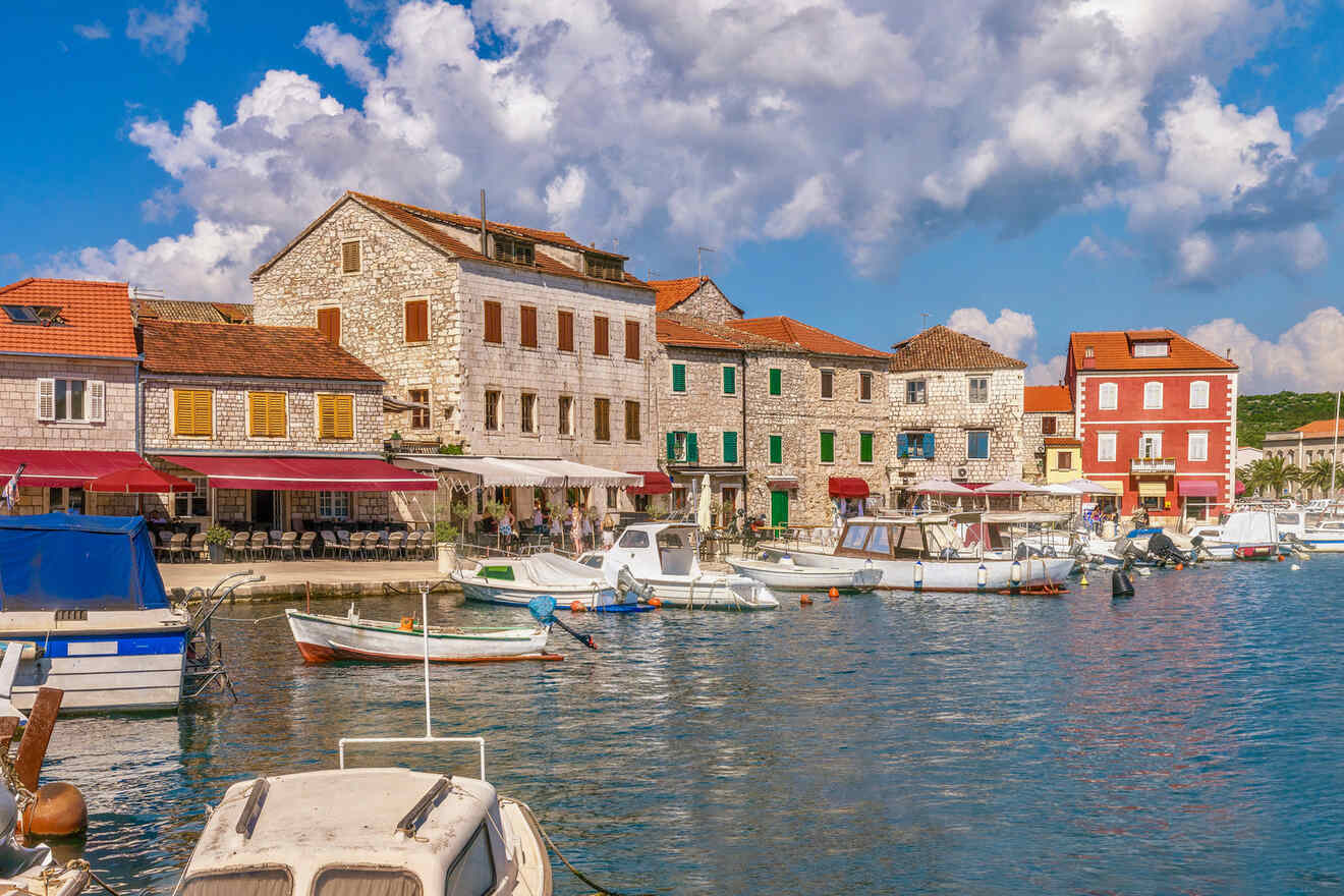 Waterfront with a row of stone buildings featuring colorful shutters, docked boats, and a blue sky with scattered clouds.